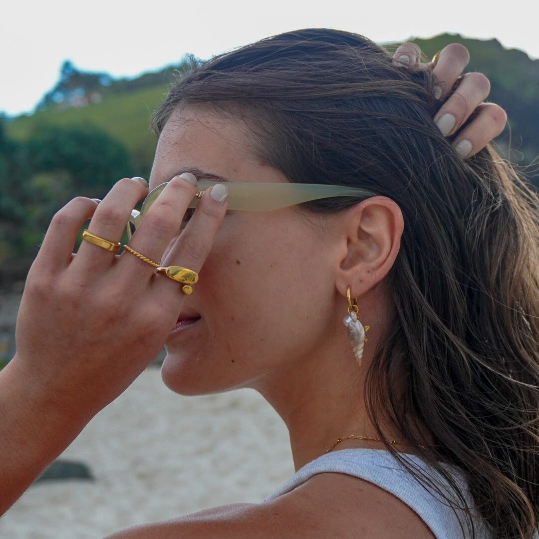 Woman shielding her eyes from the sun with a hand, wearing gold rings and earrings, with a blurred natural background.