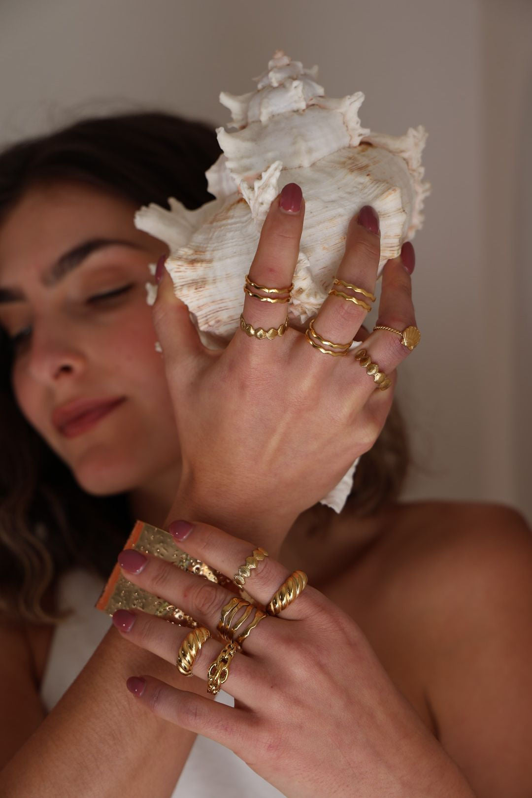 Woman holding a large shell with gold rings on her fingers against a neutral background