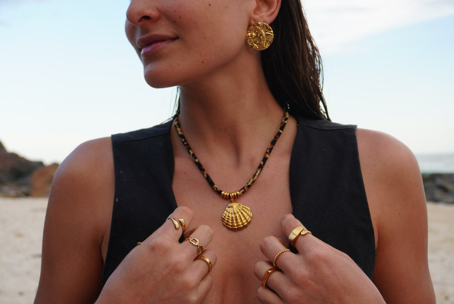 Woman wearing gold jewelry including earrings, necklace, and rings on a beach.