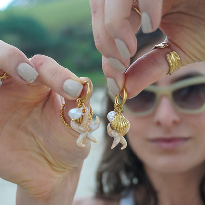 Person holding gold and shell earrings with a blurred natural background