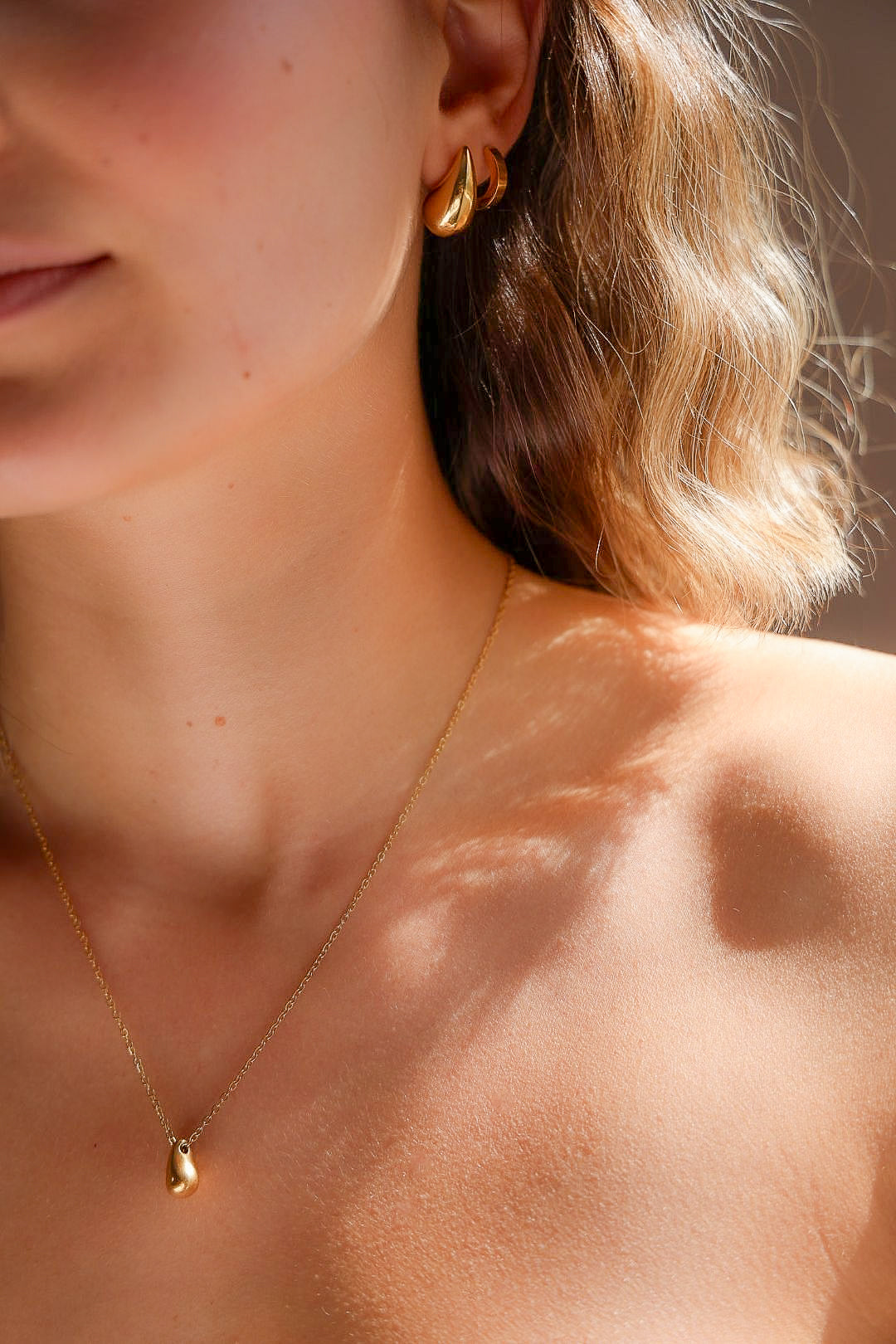 Close-up of a person wearing gold earrings and a necklace with sunlight casting shadows on skin.