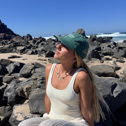 Woman sitting on rocks by the ocean wearing a white outfit and green cap.