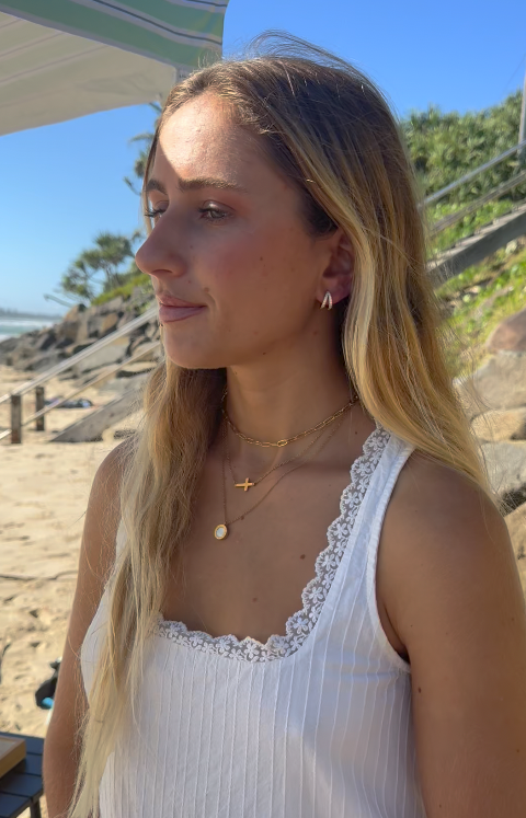 Woman with long blonde hair wearing a white sleeveless top and gold jewelry, standing on a beach.