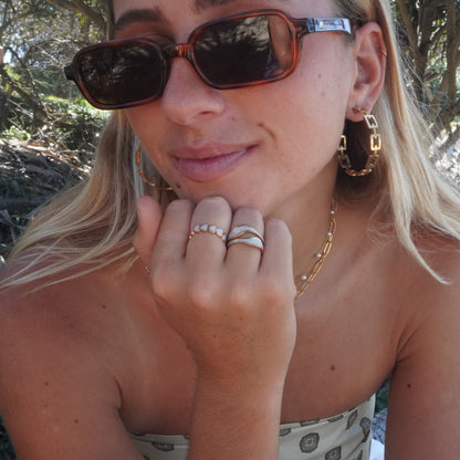 Woman wearing sunglasses and gold jewelry outdoors with trees in the background