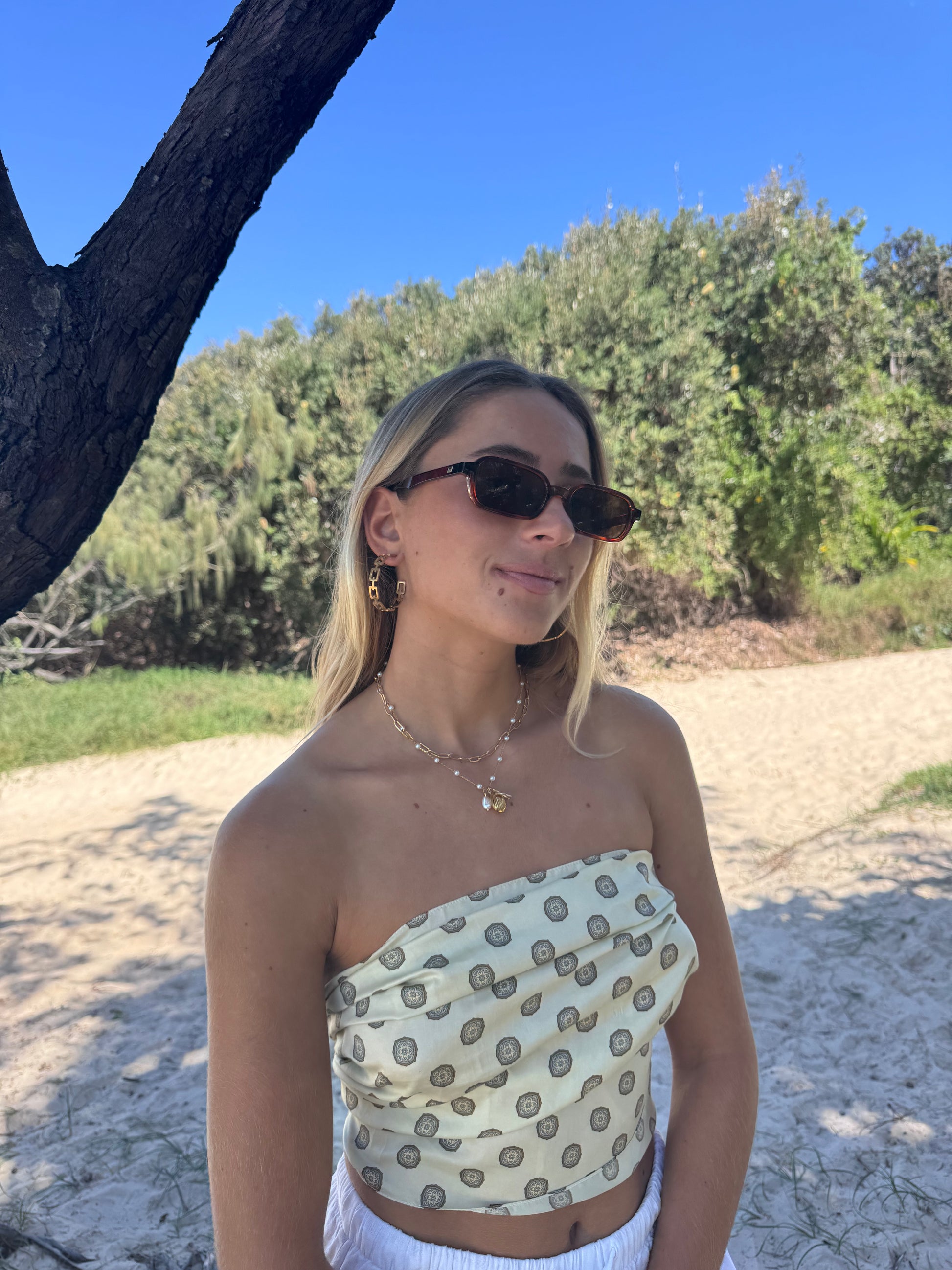 Woman wearing a strapless top with a pattern, sunglasses, and white pants standing outdoors.