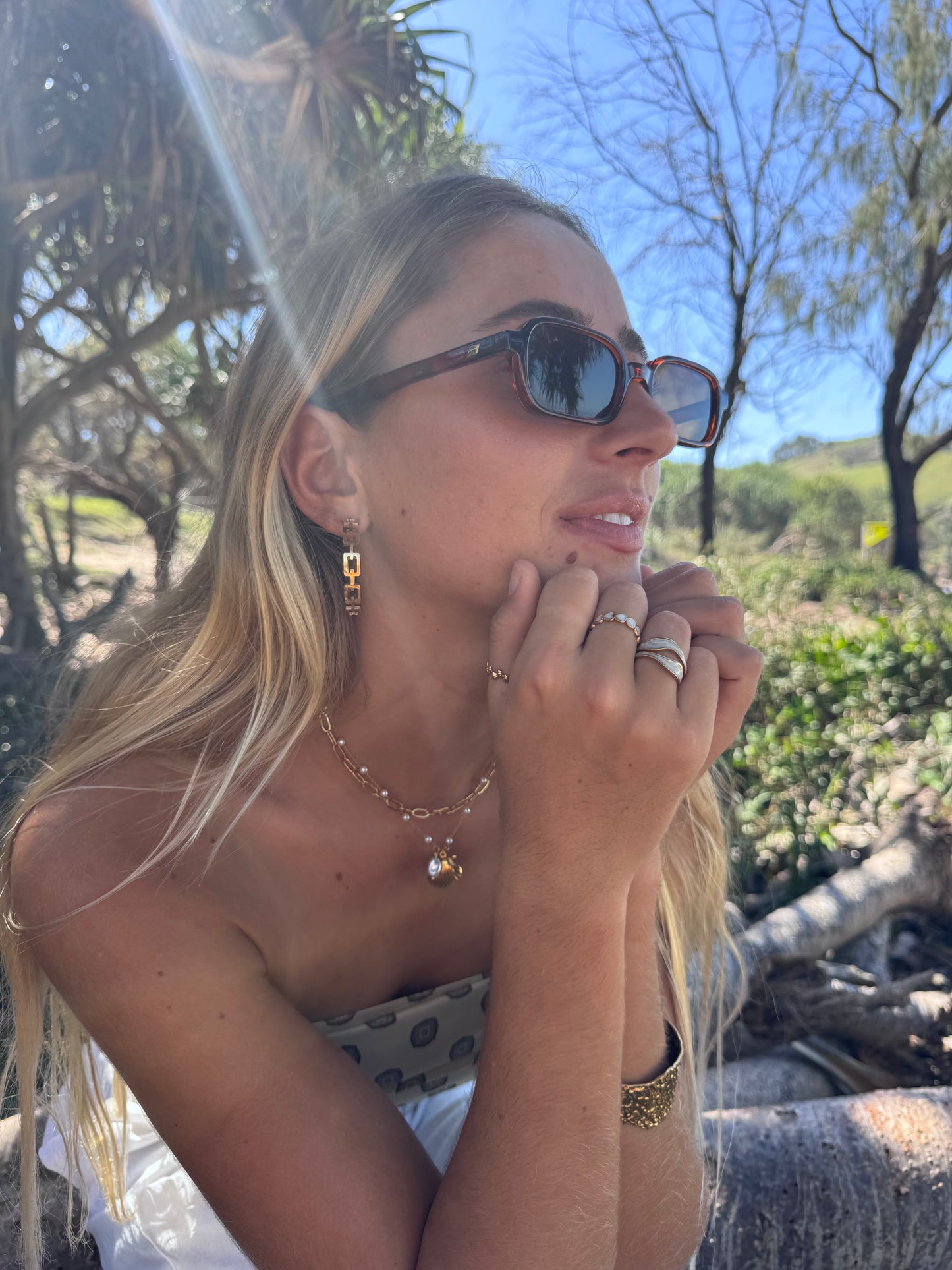 Woman wearing sunglasses and jewelry outdoors with trees and sky in the background